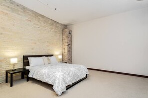 A bedroom with a brick wall, a bed with a patterned bedspread, two bedside lamps, and a small brick column. - Spacious Exchange District Condo | 1 Bed | 1 Bath | (Winnipeg)