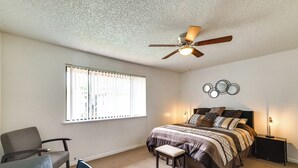 Ein Schlafzimmer mit einem Bett, einem Sessel, einem Schreibtisch, einem Deckenventilator und einem Fenster mit Jalousien.