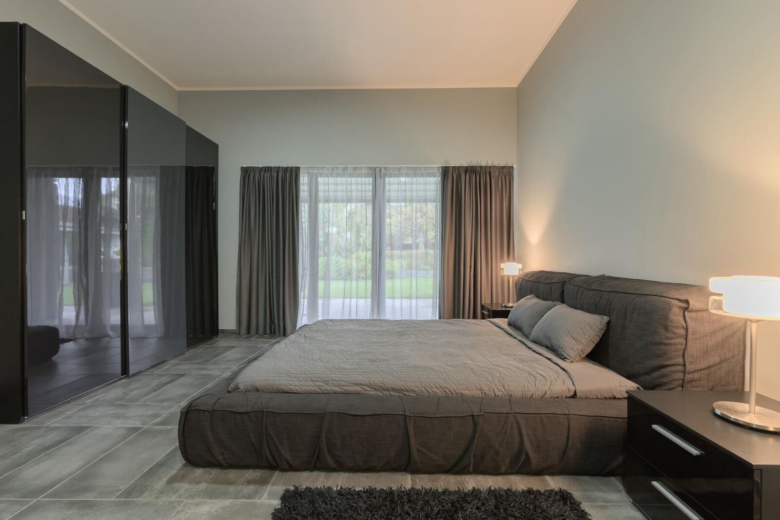 Une chambre à coucher moderne, dotée d’un grand lit, d’une table de chevet, d’une lampe et offrant une vue sur l’extérieur grâce à de grandes fenêtres.