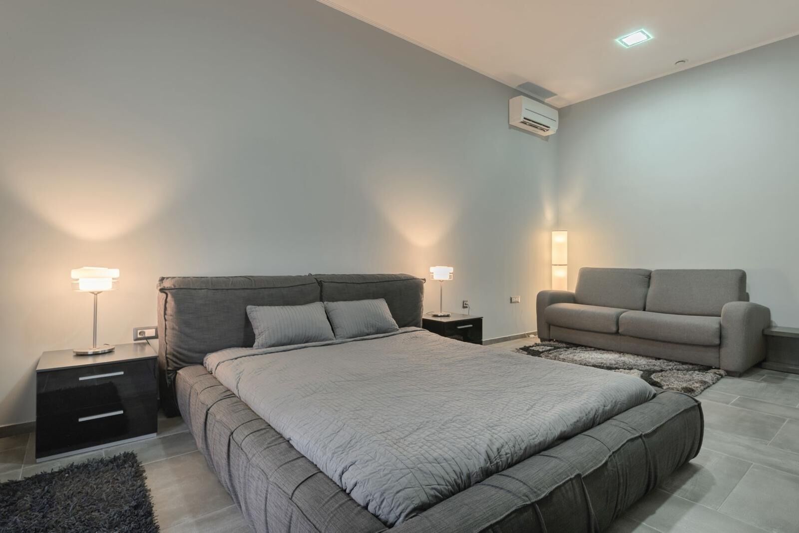 A modern bedroom with a bed, bedside tables, a sofa, and an air conditioning unit.