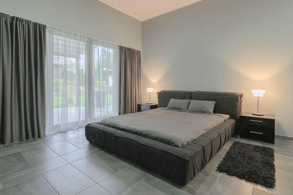 A modern bedroom with a large bed, two bedside tables, and a view through a window with curtains.