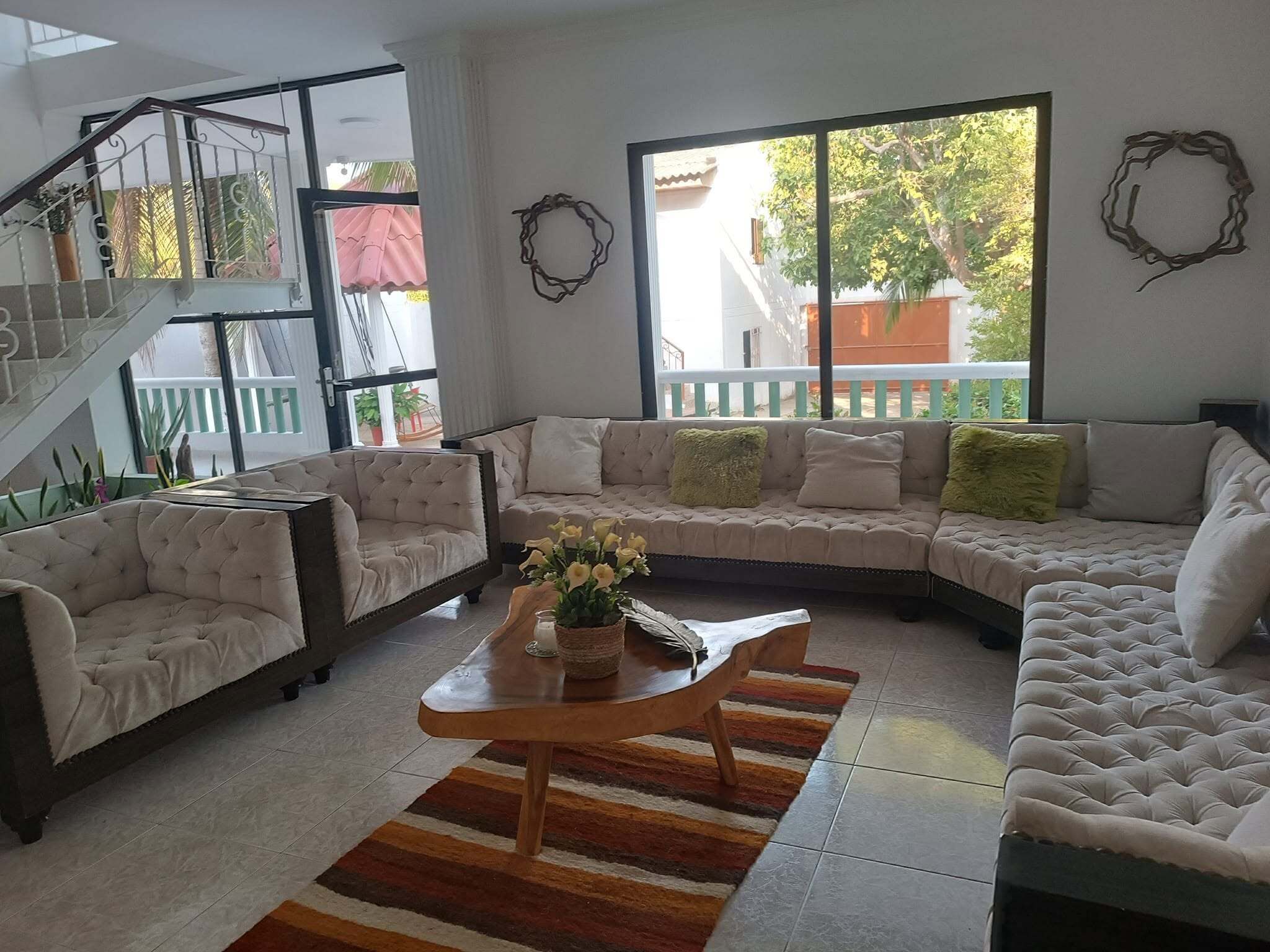Una sala moderna con un sofá seccional, dos sillones y una mesa de centro. Se observa una escalera con barandilla, un ventanal con vistas al exterior y una guirnalda decorativa en la pared.