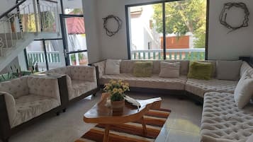 Una sala moderna con un sofá seccional, dos sillones y una mesa de centro. Se observa una escalera con barandilla, un ventanal con vistas al exterior y una guirnalda decorativa en la pared.