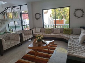 Una sala moderna con un sofá seccional, dos sillones y una mesa de centro. Se observa una escalera con barandilla, un ventanal con vistas al exterior y una guirnalda decorativa en la pared.