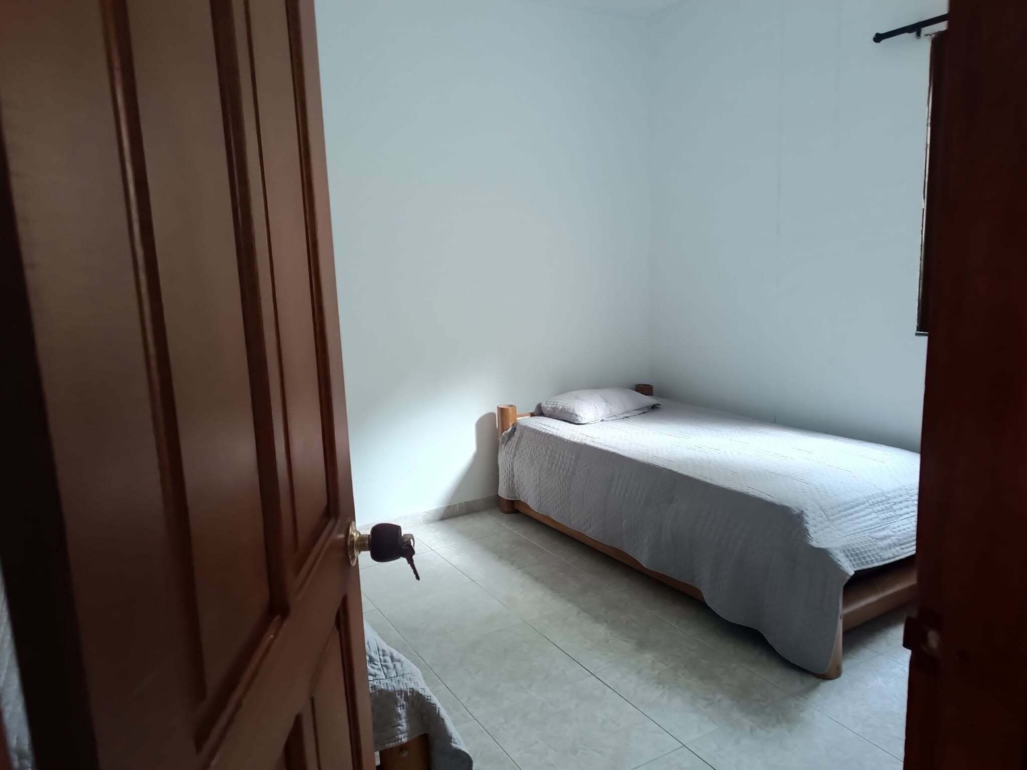 Pequeña habitación con una cama individual, puerta de madera y piso de baldosas.
