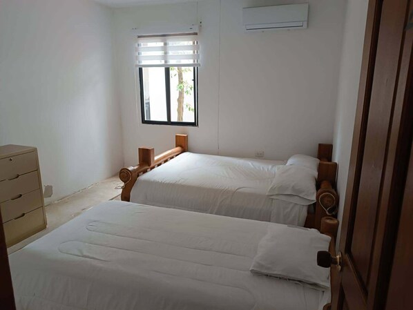 Una habitación con dos camas, una ventana con persianas, un aparador de madera y un aire acondicionado.