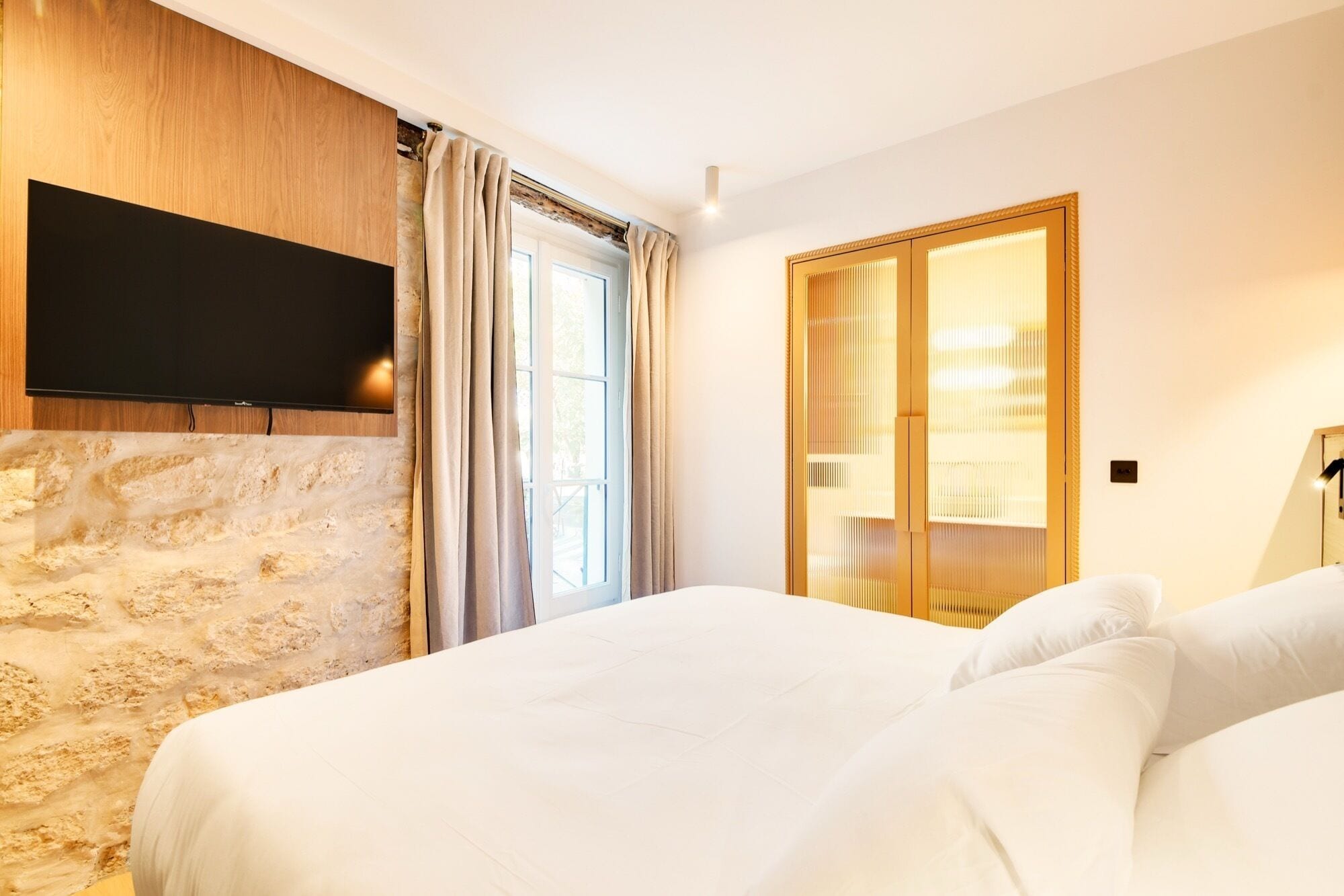 Habitación de hotel con cama, televisión montada en un panel de madera, armario y ventana con cortinas.