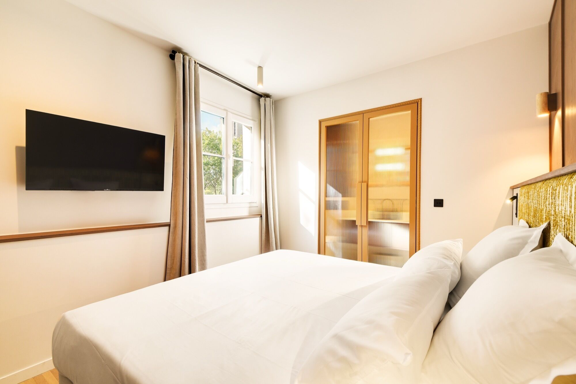 Une chambre d’hôtel comprenant un lit, un téléviseur fixé au mur, une fenêtre avec des rideaux et une armoire en bois dotée d’un miroir.