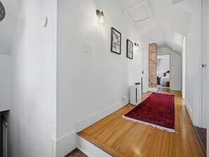 Ein Korridor mit Holzboden, weißen Wänden und einem roten Teppich. Ein Abschnitt der Wand besteht aus Ziegeln, und eine Tür führt in einen anderen Raum.