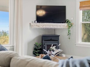 A living room with a flat-screen TV mounted on a shelf above a fireplace, a sofa, and a window with a view of trees. - The Hartson  Vintage Charm on the Ben Burr Trail (Spokane)