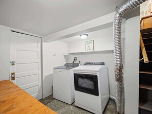 A laundry room with two washing machines, a dryer, a door, and a shelf with a framed picture. - The Hartson  Vintage Charm on the Ben Burr Trail (Spokane)