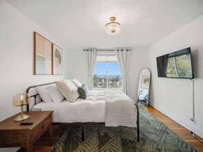Une chambre à coucher avec un lit, une table de chevet, une lampe et une vue sur la ville par la fenêtre.