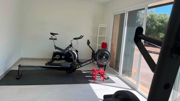 Un gimnasio en casa con una bicicleta estática, un ventilador de pie y un taburete rojo.