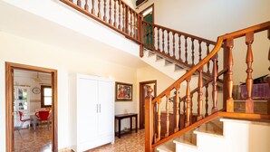 Interior de una casa de varios niveles con una escalera de madera, un comedor con sillas rojas, un armario blanco y un sofá colorido.