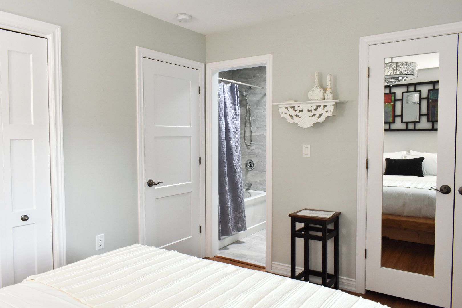 A bedroom with a bed, a door, a shower, a small table, and a mirror.