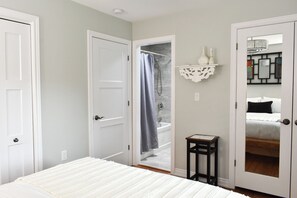 A bedroom with a bed, a door, a shower, a small table, and a mirror.