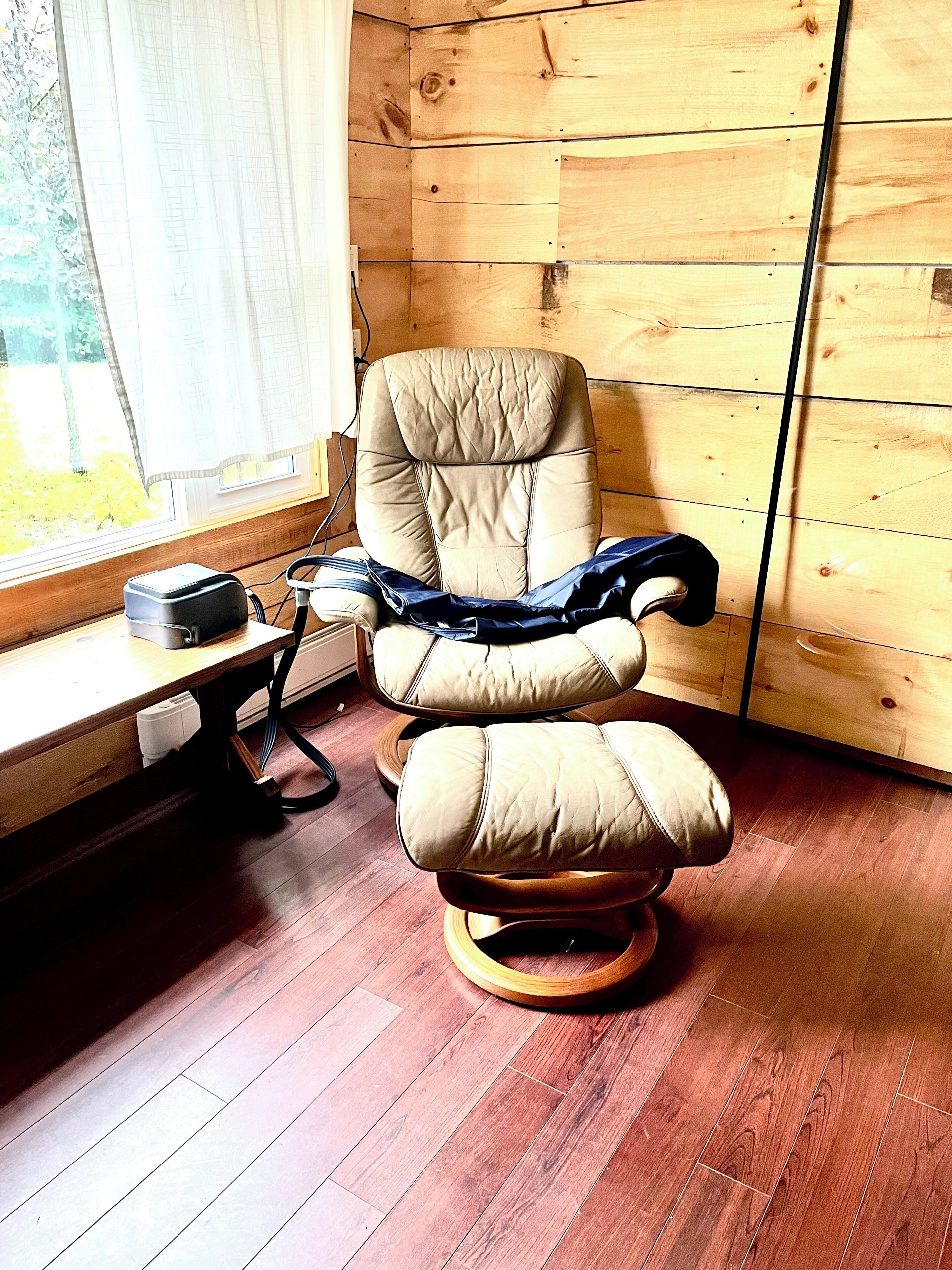 A massage chair with a footrest, positioned next to a window with a curtain, in a room with wooden walls and flooring.