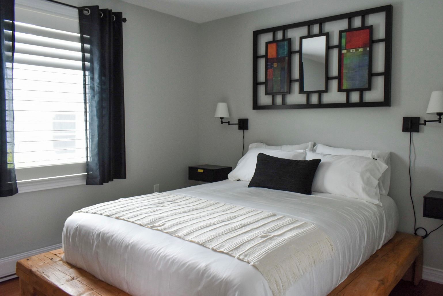 A bedroom with a bed, a nightstand, a wall-mounted lamp, and a window with curtains.