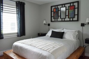 A bedroom with a bed, a nightstand, a wall-mounted lamp, and a window with curtains. - The Athlete Retreat / Sauna / Cold plunge / red light / huge kitchen (Port Colborne)