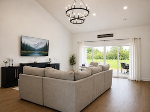 Una sala moderna con un sofá modular, un televisor de pantalla plana, una araña y un ventanal con vistas a la vegetación.