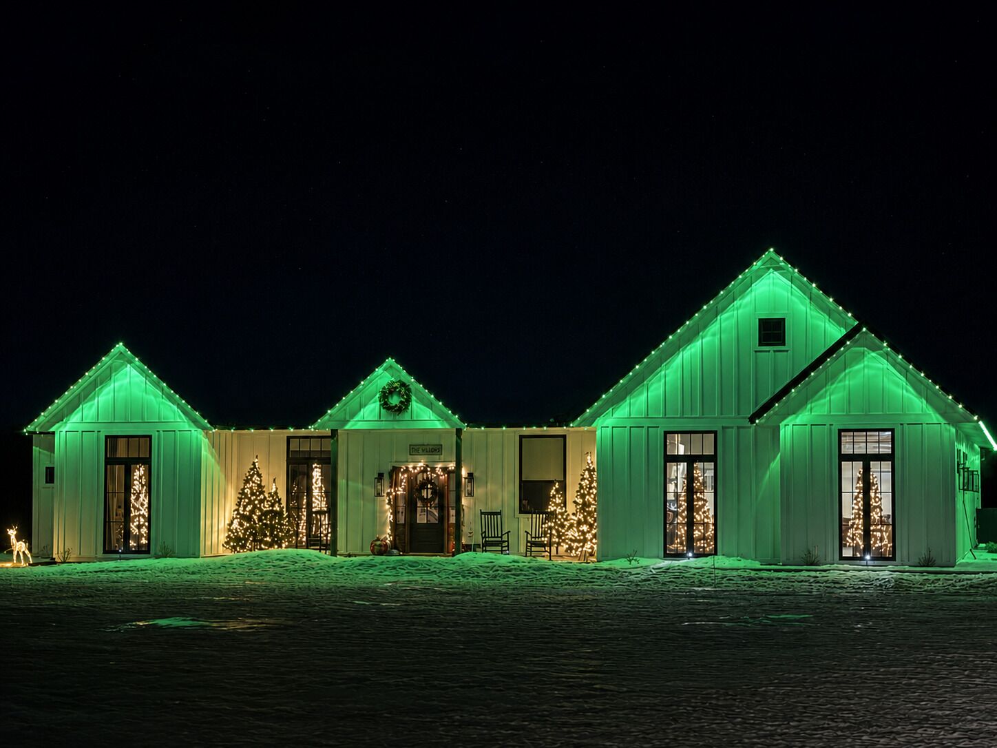 A row of three white buildings with green roofs and Christmas lights, each with a front door and windows.
