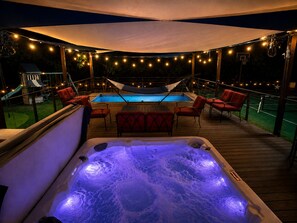 A hot tub with a wooden deck, surrounded by red chairs and a pool. - The Playhouse | Your Own Private Family Fun Resort (Bastrop)