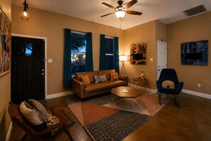 A living room with a brown leather sofa, a coffee table, a blue armchair, a floor lamp, and a ceiling fan. - The Playhouse | Your Own Private Family Fun Resort (Bastrop)