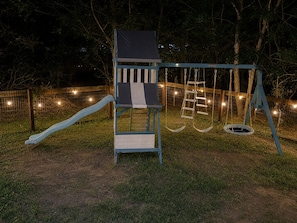A playground with a slide, a climbing structure, and swings. - The Playhouse | Your Own Private Family Fun Resort (Bastrop)