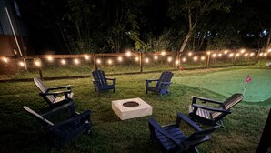 Et udendørs opholdsområde med Adirondack-stole og et bålfyr, omgivet af lyskæder.