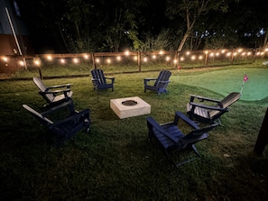 An outdoor seating area with Adirondack chairs and a fire pit, surrounded by a string of lights. - The Playhouse | Your Own Private Family Fun Resort (Bastrop)