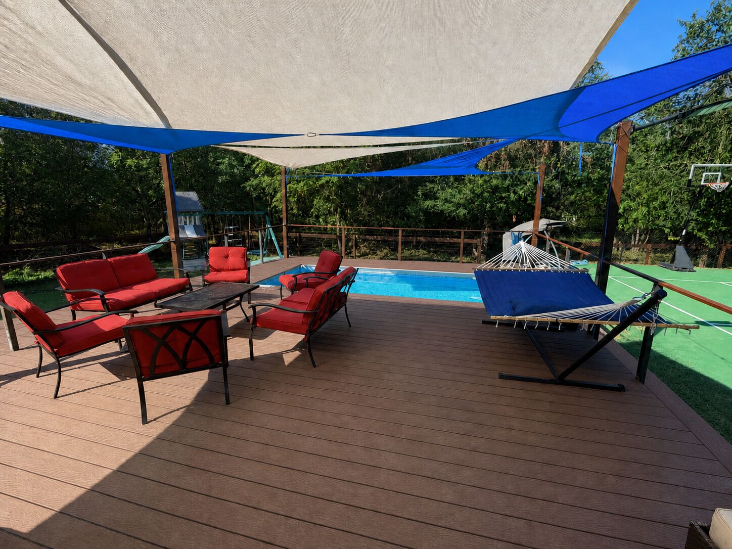 Een terras buiten met een zwembad, rode stoelen, een tafel en een hangmat.