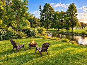 A grassy outdoor area with a fire pit and two chairs, surrounded by trees and a pond. - 3-bedroom bungalow in welcoming downtown Fenton on the Shiawassee River. (Fenton)