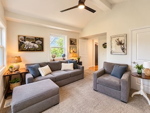 A spacious living room with a grey sectional sofa, a grey armchair, and a matching ottoman. There are two side tables with lamps and decorative items. The room has a high ceiling with a ceiling fan, large windows, and a door leading to another room. - 3-bedroom bungalow in welcoming downtown Fenton on the Shiawassee River. (Fenton)