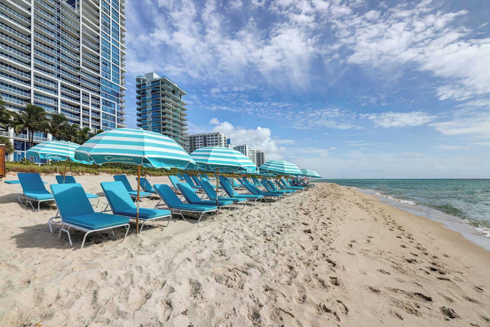 A beach with rows of blue lounge chairs and striped umbrellas, a clear sky, and a calm sea.