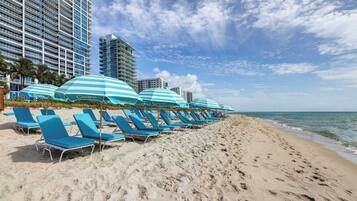 A beach with rows of blue lounge chairs and striped umbrellas, a clear sky, and a calm sea.