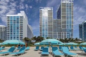 A beachfront hotel with multiple high-rise buildings, a sandy beach with lounge chairs and umbrellas, and palm trees.