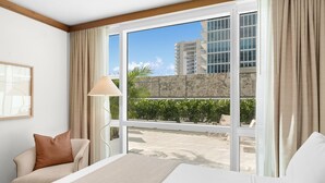 Ett hotellrum med en säng, en soffa, en lampa och ett stort fönster med utsikt över en byggnad och grönområden utanför.