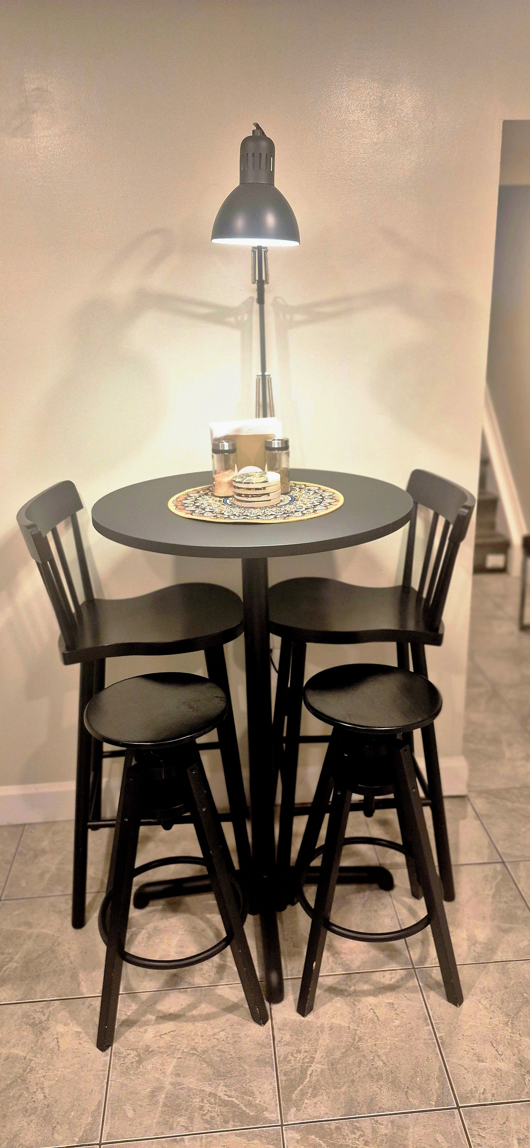 A round dining table with four chairs, a table lamp, and salt and pepper shakers on a decorative coaster.