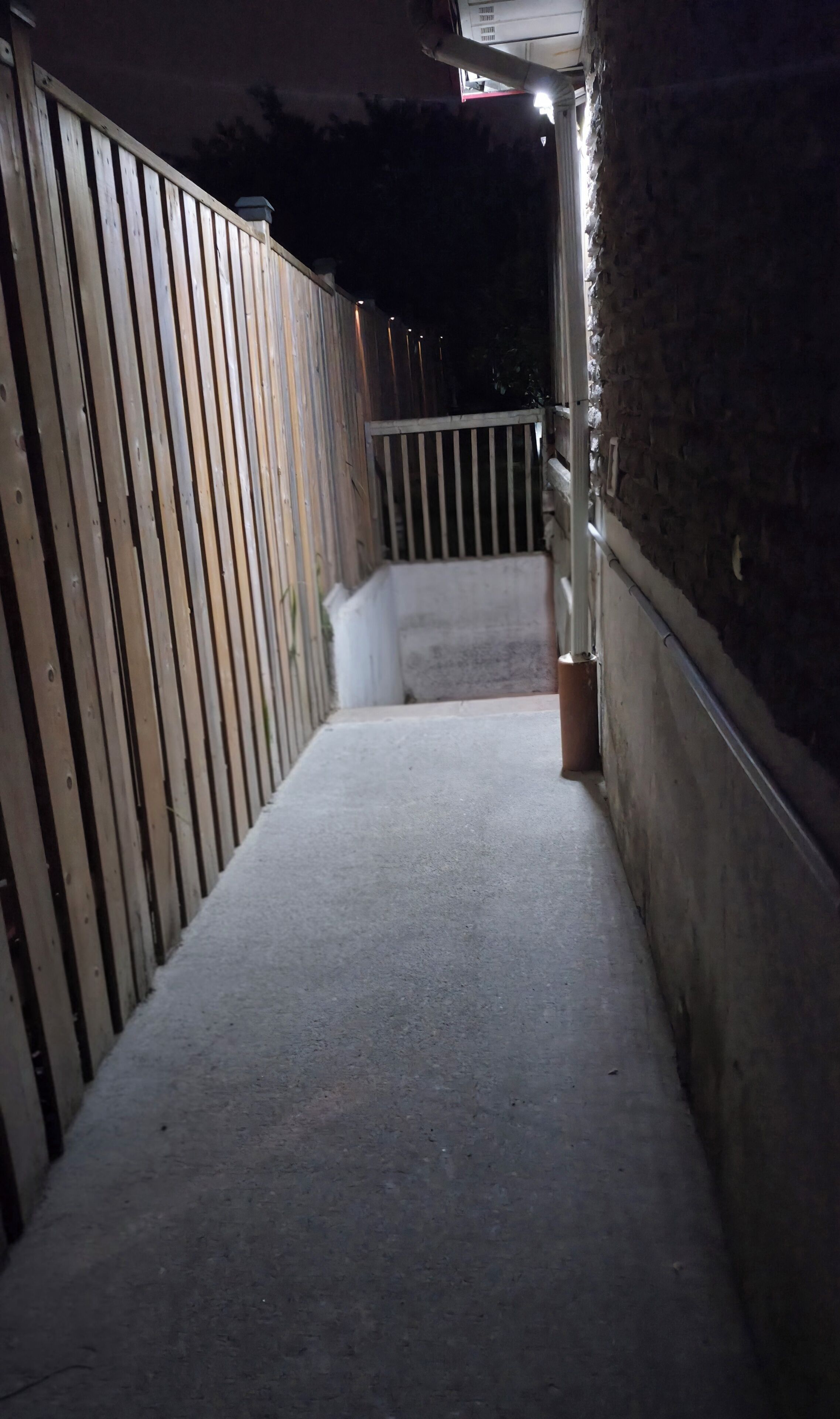 A narrow concrete pathway leading to a gated area with a wooden fence on one side and a stone wall on the other.