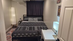 A compact bedroom with a bed, bedside table, lamp, and a desk with a chair.
