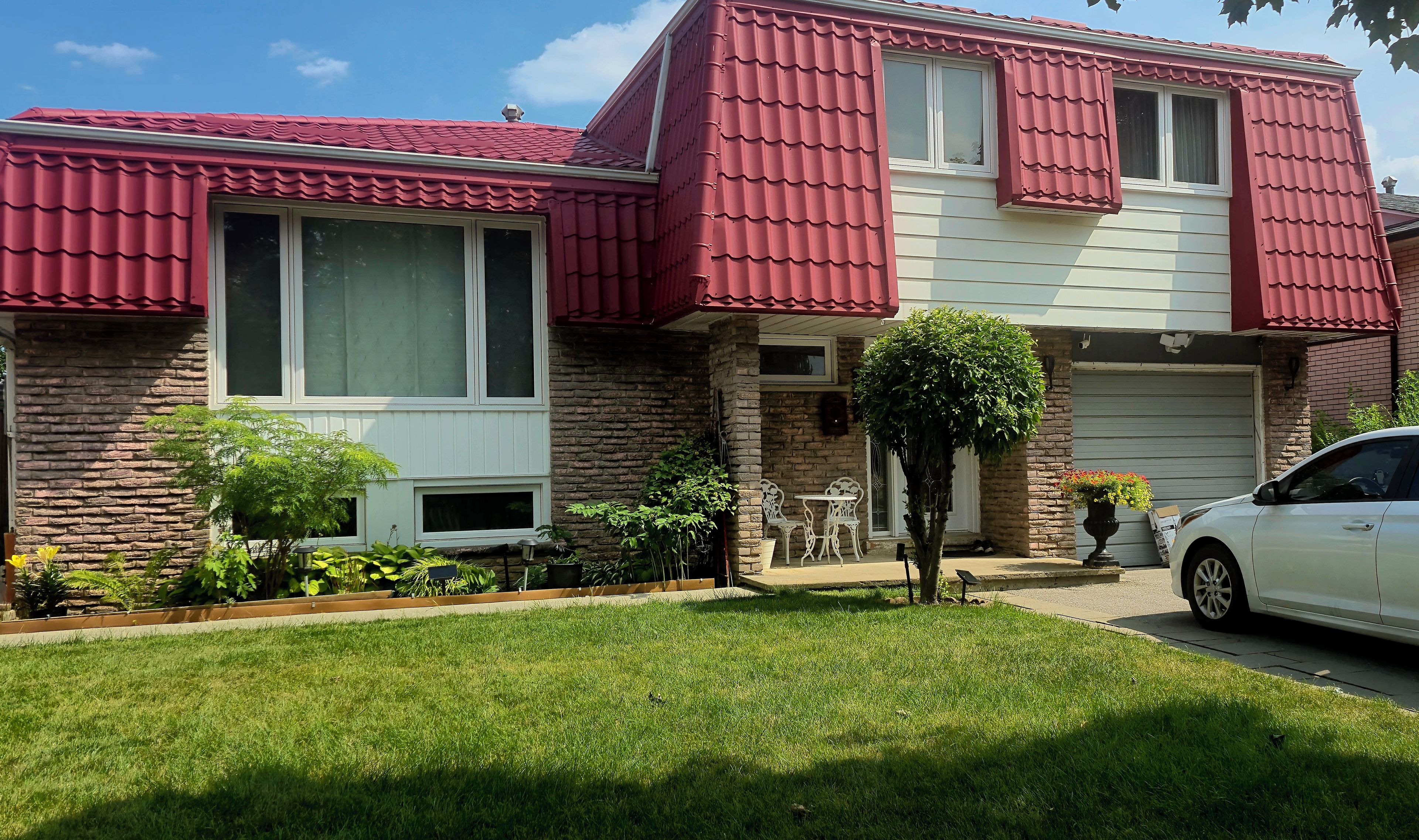 A two-story house with a brick facade, red roof, and a front yard with a lawn and potted plants.
