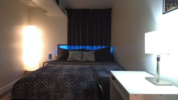 A hotel room with a bed, a desk, a lamp, a nightstand, and a wall-mounted picture.