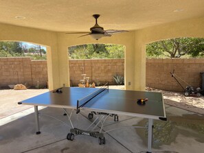 Mesa de ping pong al aire libre con un ventilador de techo, frente a un muro de ladrillo y follaje.
