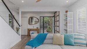Una sala de estar moderna con un sofá, una mesa de centro y un espejo. Se ve una escalera a la izquierda y una puerta corrediza de cristal que da a un balcón.