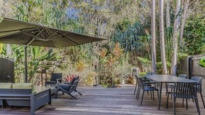 Un patio al aire libre con una mesa y sillas para comer, rodeado de abundante vegetación y una terraza de madera.