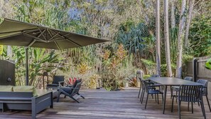 An outdoor patio with a dining table and chairs, surrounded by lush vegetation and a wooden deck. - Samsara in Suffolk Park (Suffolk Park)