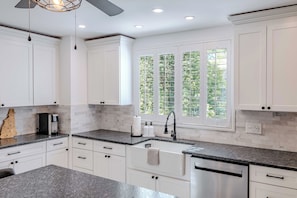 A modern kitchen with white cabinetry, a granite countertop, a built-in dishwasher, and a window with a view of greenery. - Modernized Historic Mountain Getaway (Littleton)