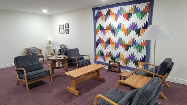 A room with a wooden coffee table, two armchairs, a small table with a lamp, and a colorful quilt on the wall.