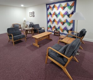 Ein Zimmer mit einem Holztisch, zwei Sesseln, einem kleinen Tisch mit Lampe und einer farbenfrohen Stegdecke an der Wand.