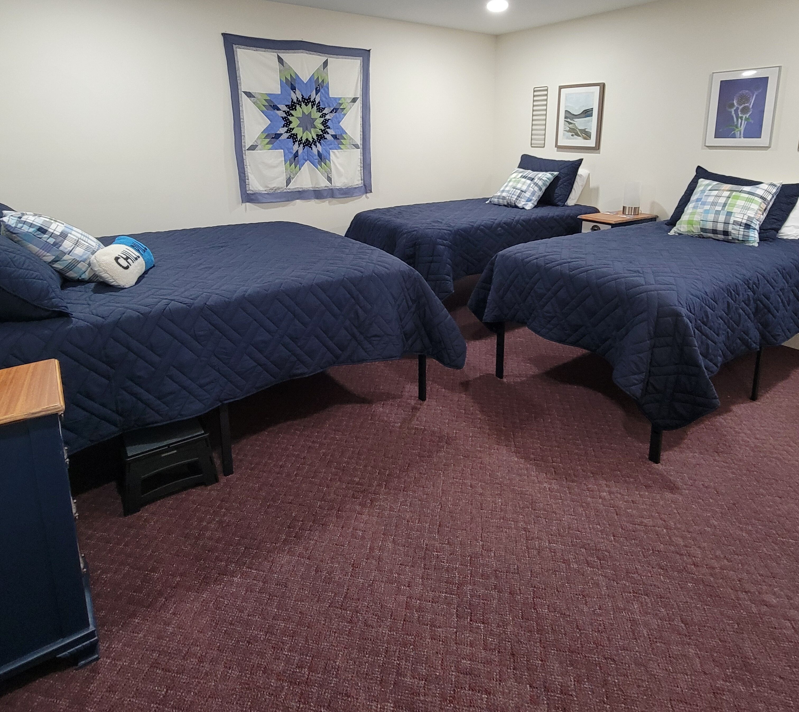 A room with three beds, each with a blue quilted bedspread and pillows, a red carpet, and wall art including a star pattern and two framed pictures.